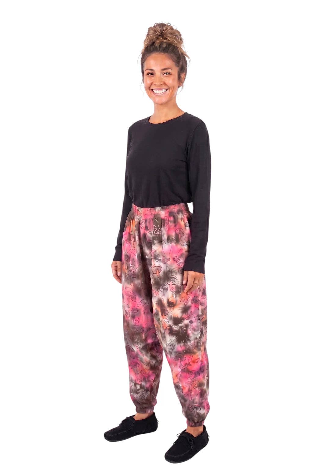 A woman modeling a pair of relaxed-fit jogger pants with a pink, red, and brown tie-dye pattern, paired with a black long-sleeve shirt and black slip-on shoes, against a plain white background.
