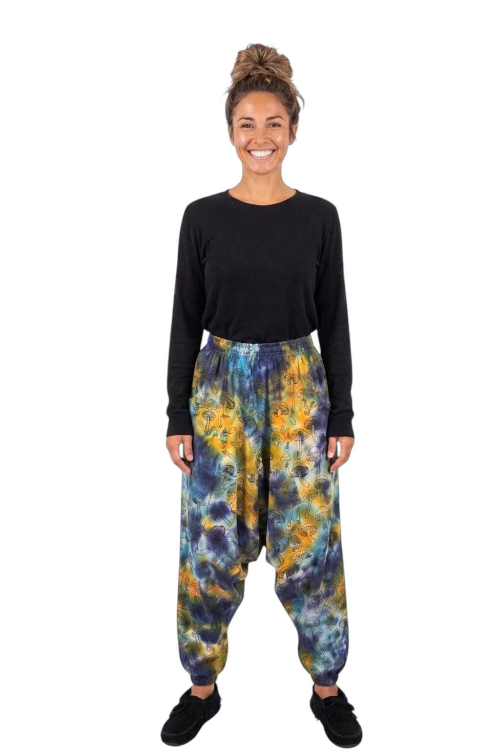 A woman modeling loose harem-style pants with a blue, yellow, and orange tie-dye pattern, paired with a black long-sleeve top and black shoes, against a plain white background.